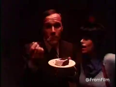 In the 16mm still from the 1970s Betty Crocker Ring Cake (Bundt cake) commercial, a man holding a plate with a slice of Bundt cake appears surprised or delighted, as he brings a forkful closer to his mouth. Beside him, a woman appears eager or excited, likely encouraging him to try the cake. The scene is set in dim lighting, creating a warm, nostalgic atmosphere typical of the era. The focus on their expressions captures the joy of sharing and enjoying homemade treats.