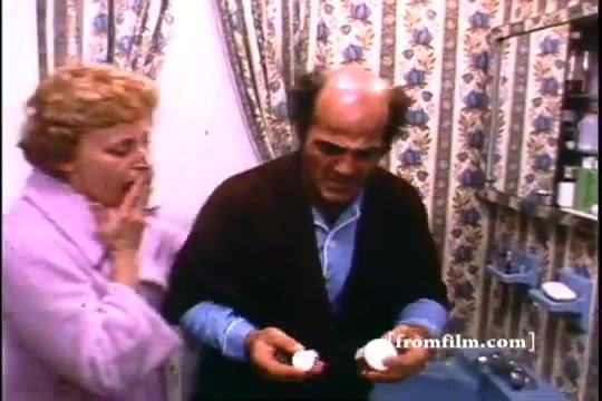 The still depicts a tense moment in a bathroom, featuring two characters: a man and a woman. The man, looking distressed, holds two bottles, while the woman appears shocked with one hand over her mouth. The decor is distinctly 1970s, with floral wallpaper and a cluttered bathroom setting, emphasizing the importance of the message about disposing of unused medicine.