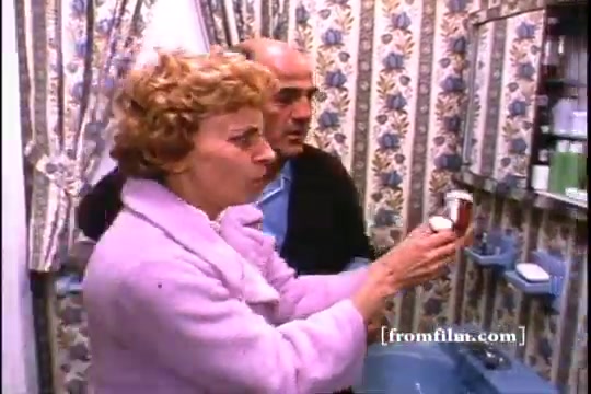 The still features a woman in a pink coat holding a medicine bottle while looking concerned, alongside a man in a black sweater. They are in a bathroom with floral wallpaper, suggesting an older setting typical of the 1970s. The scene emphasizes the theme of disposing of unused medicine, reflecting public health messaging from that era.