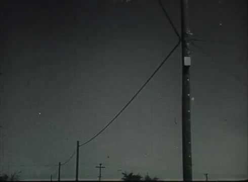 The still from 'Bell System Film - Drop Wire Runs Part 1 (a)' shows a telephone pole with wires extending towards the viewer, symbolizing the infrastructure that delivers telephone service. The background features a gradient sky, emphasizing the pole and wires as key elements. This image reflects the late 1940s to early 1950s context of telephone installation and the connection between central offices and customers.