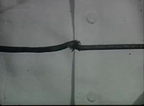 The still from 'Bell System Film - Drop Wire Runs Part 1 (a)' shows two lengths of telephone wire making contact with each other, possibly indicating a repair or connection point. The image highlights a close-up of the wires, emphasizing their condition and the technique used to join them. The context suggests it's part of a tutorial on telephone service infrastructure from the late 1940s to early 1950s, focusing on practical aspects of installation.