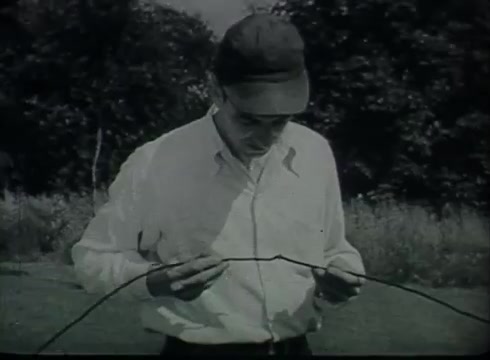 The still from 'Bell System Film - Drop Wire Runs Part 1 (a)' depicts a male technician wearing a cap and a white shirt, inspecting a length of drop wire. He stands outdoors, surrounded by greenery, indicating a practical demonstration of telephone wire installation. The film illustrates the process of connecting telephone lines from utility poles to customer premises, typical of the informational style used in the late 1940s to early 1950s.