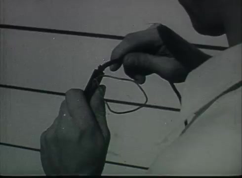 The still from the Bell System film 'Drop Wire Runs Part 1 (a)' depicts a close-up of a person’s hands as they handle a telephone wire. The image focuses on the technique of connecting or preparing the wire, likely showcasing the steps involved in the installation process. The historical context suggests this film serves an educational purpose, illustrating telecommunications practices from the late 1940s to early 1950s.