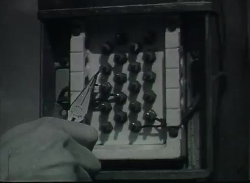 The still shows a close-up of a utility box, likely part of a telephone system, with a hand using pliers to manipulate connections. The box features multiple terminals and is set in a muted, monochrome style typical of the late 1940s to early 1950s. This moment illustrates the technical process of connecting drop wires from overhead poles to customer lines, emphasizing the practical aspects of telephone installation.