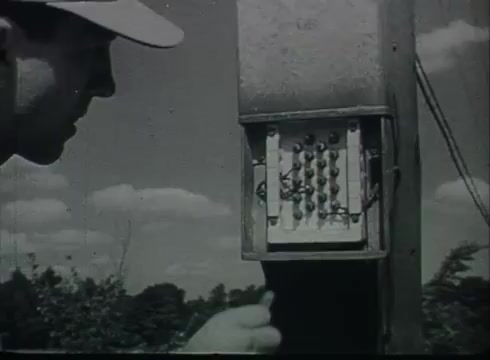 The still depicts a technician working on a telephone junction box mounted on a utility pole. He is wearing a cap and is focused on the internal wiring of the box, which contains multiple connections. The setting features a clear sky with some clouds in the background, indicating a daytime scene. This image illustrates the process of connecting telephone wires from the central office to the customer, highlighting the hands-on nature of telephone service installation in the late 1940s to early 1950s.