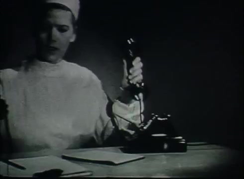 The still features a woman, likely a telephone operator, wearing a white uniform and a cap, seated at a desk. She is holding a black telephone receiver with one hand while looking down at some notes on the table. The image captures a moment of communication, emphasizing the operational aspect of telephone service during the late 1940s or early 1950s, likely illustrating the connection process between the Central Office and customers. The overall aesthetic reflects the film's educational focus on telephone infrastructure.