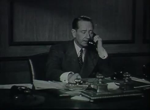 The still depicts a middle-aged man in a suit seated at a desk, actively engaged in a telephone conversation. He appears focused, with papers and office supplies scattered on the desk. The setting suggests a professional environment, likely in a telecommunications context given the film's subject matter. The lighting is subdued, characteristic of the era, contributing to a serious atmosphere suitable for a corporate setting.