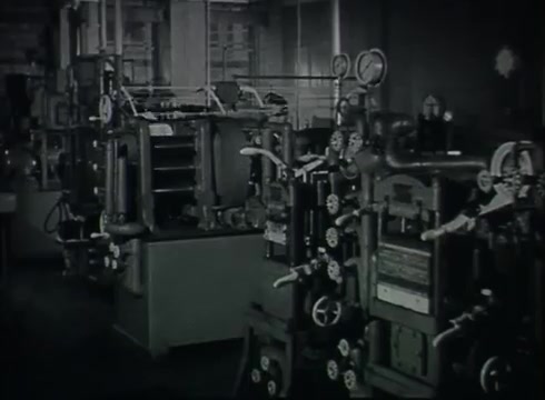 The still from 'Bell System Film - Drop Wire Runs Part 1 (a)' depicts a Central Office environment from the late 1940s to early 1950s. The image shows various machines and equipment, including panels with gauges, levers, and valves, all housed within a well-lit industrial space. This setting highlights the technological processes involved in telephone service delivery, illustrating the complexity of the infrastructure supporting communication networks during that era.