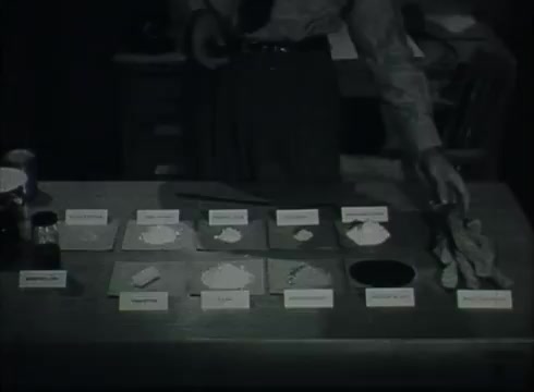 The still features a table displaying labeled samples of various materials related to telephone wire installation, such as insulation and connectors. A person, likely a technician or instructor, is positioning one of the materials, indicating a practical demonstration. The setting appears to be a Central Office environment, typical of the late 1940s to early 1950s, emphasizing hands-on training in telecommunications.