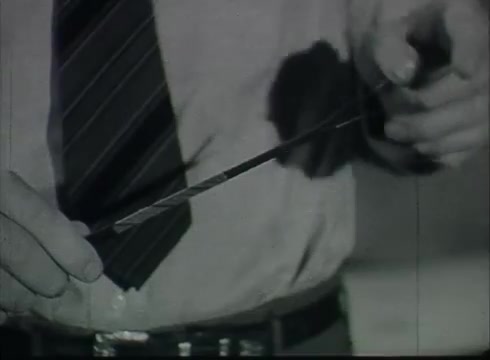 The still captures a person, dressed in a collared shirt and tie, demonstrating a piece of telephone wire, possibly indicating its construction or application. The image likely focuses on the process of connecting drop wires from telephone poles to customer premises, a key topic in the Bell System film from the late 1940s or early 1950s. The hand positioning suggests a hands-on instructional element, characteristic of technical training visuals from that era. The film aims to educate viewers about telecommunications infrastructure.