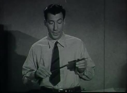 The still features a man in a collared shirt and tie, speaking while holding a small tool or instrument, likely related to telephone installation. He stands in front of a simple background, indicative of an educational or instructional setting typical of the late 1940s to early 1950s. The film focuses on the processes involved in connecting telephone wires from poles to customers, highlighting practical aspects of telecommunications technology.