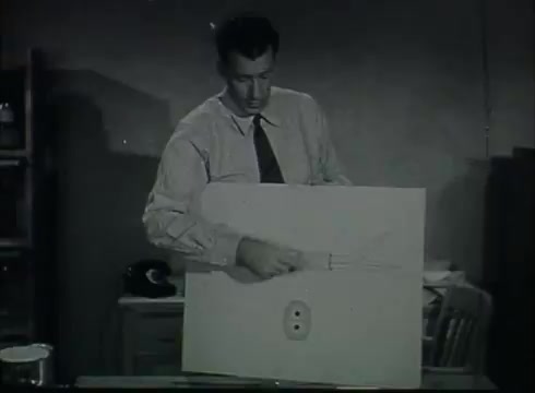 The still features a man dressed in a light-colored shirt and tie, demonstrating a telephone installation process. He is holding a model or illustration on a board, likely depicting the connection between telephone wire and a wall outlet or similar fixture. The setting appears to be an office or workshop, reflecting the educational nature of the film. This scene is part of a Bell System film from the late 1940s to early 1950s, designed to explain telephone wire installations to viewers.