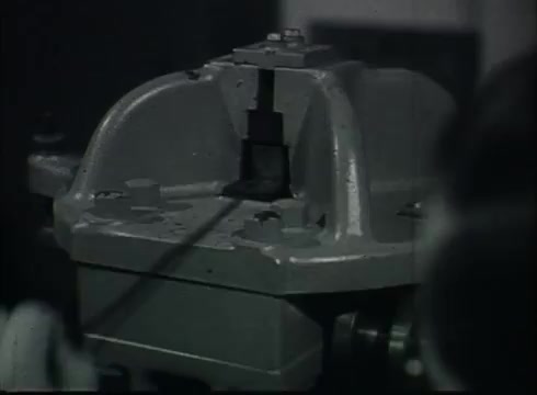 The still depicts a piece of industrial equipment used in the process of preparing telephone wire, likely within a Central Office setting. The focus is on a metallic apparatus that appears to be a clamp or machine component, holding a length of wire, showcasing the mechanics involved in connecting telephone lines from poles to customers. The overall style reflects the mid-20th-century aesthetic typical of instructional films from that era.