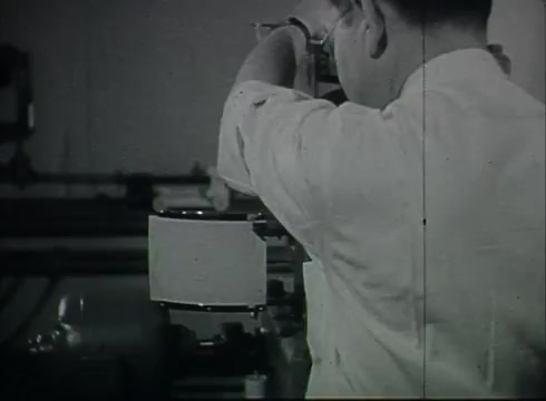 The still from 'Bell System Film - Drop Wire Runs Part 1 (a)' showcases a technician in a white shirt focused on manipulating equipment within a Central Office setting. The background suggests a technical environment, likely related to telecommunications, illustrating the processes involved in connecting telephone wires from utility poles to customers. The image captures the era's attention to detail and the importance of precise operations in telephone service provision.
