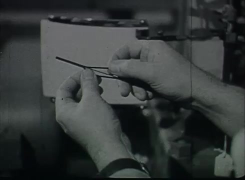 The still from 'Bell System Film - Drop Wire Runs Part 1 (a)' shows a person's hands holding a piece of wire. The background appears to be part of a Central Office environment, indicative of the late 1940s to early 1950s period. The focus on the wire suggests the film's instructional theme, likely detailing the process of connecting telephone wires from utility poles to customer premises.