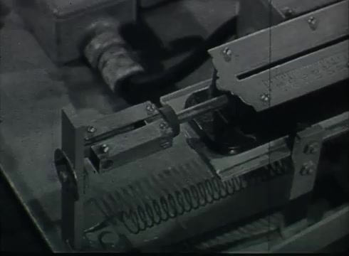 The still from 'Bell System Film - Drop Wire Runs Part 1 (a)' shows a close-up of a mechanical device, likely part of the Central Office equipment used during the late 1940s to early 1950s. It features metal components, including a spring mechanism, indicating its function in telecommunications. The clear focus on machinery highlights the technical processes involved in connecting telephone wire from utility poles to customer premises.