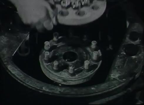 The still shows a close-up of a mechanical device, likely related to the processing or installation of telephone wire. A hand is seen manipulating a component above a circular piece with multiple bolts. This scene captures a moment in the Central Office, illustrating the equipment and tools used in telephone wire management during the late 1940s to early 1950s.