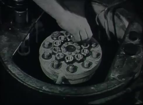 The still depicts a close-up of a hand manipulating a circular component, possibly a terminal or connector, within a Central Office setting. The environment appears industrial and utilitarian, typical of the late 1940s to early 1950s. This scene likely illustrates the process of connecting telephone wires as part of the telecommunication infrastructure, emphasizing the technical aspects of telephone service setup.