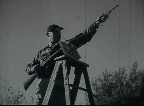 The still from 'Bell System Film - Drop Wire Runs Part 1 (a)' features a man standing on a ladder with a telephone pole in the background. He is wearing a cap and appears to be holding a tool or equipment, possibly for installation or maintenance purposes. The scene likely illustrates a step in the process of connecting telephone wires from poles to customers, typical of the film's instructional content from the late 1940s to early 1950s. The black-and-white imagery evokes the era's documentary style, emphasizing telecommunication work.