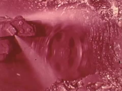 The still from the 'Mining Enforcement and Safety Administration PSA' depicts a close-up of mining machinery in action, likely showing a rotating wheel or cutter as it moves through rock or soil. The image has a reddish tint, emphasizing the environment of a mining site and the equipment's function in extracting resources. The focus on the machinery highlights the importance of safety and enforcement in mining operations.