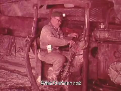 The still from the 'Mining Enforcement and Safety Administration PSA' shows a miner operating machinery in a mine setting. He appears focused and responsible, showcasing the importance of safety and enforcement in mining operations. The scene emphasizes the working conditions and practices involved in the mining industry.