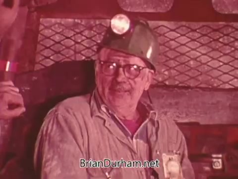 The still features an older male miner wearing a hard hat with a lamp, sitting in a mine. He expresses a serious demeanor, likely discussing safety concerns. The background suggests a typical mining environment, emphasizing the focus on mining safety and regulations. The image captures the essence of the Mining Enforcement and Safety Administration PSA.