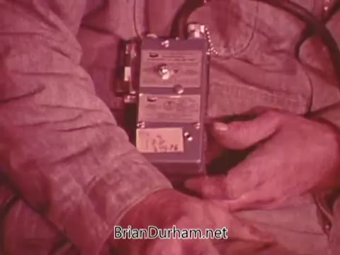 The still features a close-up of a miner's hands holding a safety device, likely a carbon monoxide detector or monitoring equipment, essential for ensuring safety in mining operations. The miner is wearing a denim jacket, suggesting a rugged work environment. The image has a vintage quality, indicative of a mid-20th century public service announcement aimed at promoting safety awareness in the mining industry.