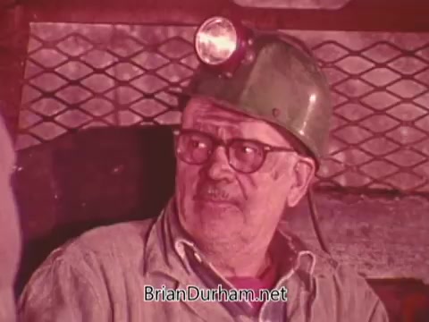 The still features an older miner wearing a helmet with a light attached, sitting in a dimly lit environment. He has glasses and a thoughtful expression, possibly engaged in conversation or reflecting. The background suggests a mining setting, emphasizing themes related to occupational safety and the mining industry. This imagery likely serves to highlight the importance of safety measures in mining.