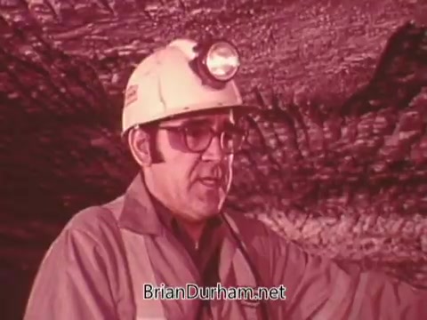 The still from 'Mining Enforcement and Safety Administration PSA' features a miner wearing a safety helmet equipped with a headlamp. He appears to be speaking or instructing, likely emphasizing safety measures in a mining environment. The background showcases the cave-like structure of a mine, emphasizing the context of mining safety. The image's color tone has a reddish hue, contributing to a serious and focused atmosphere.