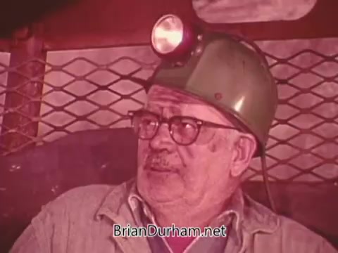 The still features an older man wearing a green mining helmet equipped with a headlamp. He has spectacles and appears to be engaged in conversation or deep thought. The background suggests an underground mining environment, emphasizing the theme of safety in mining. The image captures a sense of seriousness regarding mining safety regulations.