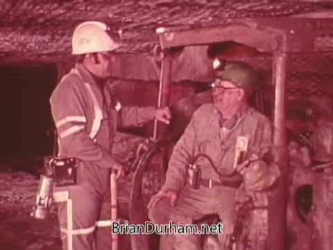 The still from the PSA features two miners in an underground setting. One miner, wearing a helmet and work attire, is standing while conversing with another seated miner, who also wears a helmet and protective gear. The scene emphasizes safety and communication in mining operations. The background suggests a typical mining environment, highlighting the importance of enforcing safety protocols in such settings.