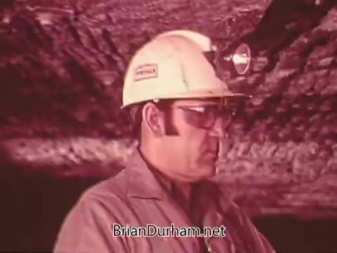 The still from the 'Mining Enforcement and Safety Administration PSA' features a miner wearing a hard hat with a headlamp and protective glasses. He appears focused and is likely in a subterranean mining environment, indicated by the rocky ceiling above him. The overall tone suggests an emphasis on safety and the working conditions in mining.
