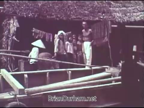 The still from the 1969 'Christian Children's Fund' PSA depicts a group of children and adults near a simple, rural dwelling. The scene reflects a community environment, showcasing a woman in a traditional conical hat and several children observing from a wooden structure. The imagery evokes themes of childhood, poverty, and the outreach efforts of the organization during that era.