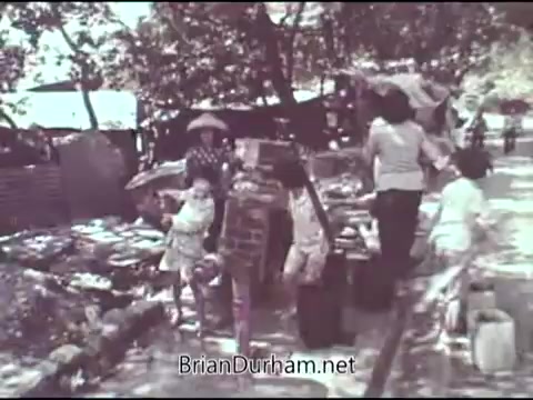 The still depicts a group of children and adults in a rural setting, likely part of a community illustrating hardship. Some individuals are engaged in daily activities, while children are prominently featured, emphasizing innocence amidst the surrounding challenges. The scene reflects themes of poverty and community support, consistent with the mission of the Christian Children's Fund in the late 1960s. The overall tone is somber yet hopeful, aiming to raise awareness and encourage assistance for those in need.