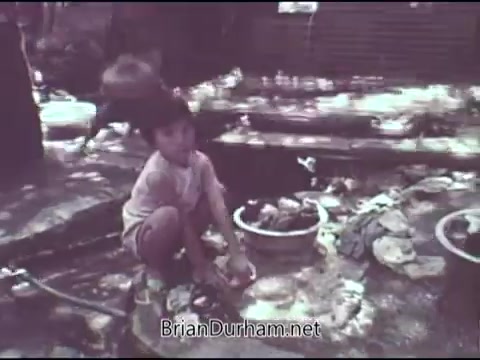 The still depicts a young child squatting on the ground amidst a cluttered outdoor setting, surrounded by various items and containers. The scene reflects a moment of everyday life, likely highlighting the struggles faced by children in impoverished conditions. The visual style is indicative of the time, with a faded color palette typical of 1969 film. This PSA aims to raise awareness about the challenges faced by children and promote support from organizations like the Christian Children's Fund.