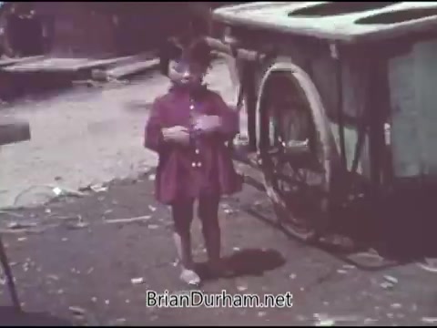 The still from the 1969 PSA for the Christian Children's Fund features a young girl wearing a pink coat standing in a gritty, outdoor setting. She appears to be in a somewhat impoverished environment, with a cart visible in the background, suggesting themes of innocence and the need for support in less fortunate circumstances. The visual style reflects the era, with muted colors typical of 16mm film.