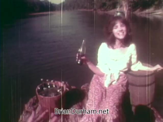 The still from the 1973 Coca-Cola nostalgia commercial depicts a woman sitting by a river, smiling and holding a Coke bottle. She is dressed in casual, colorful clothing with a bohemian style. A basket filled with Coke bottles is beside her, emphasizing the carefree, joyful theme of enjoying Coke in a natural setting. The overall atmosphere is warm and nostalgic, capturing a sense of simplicity and happiness.