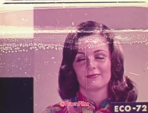 The still depicts a woman with closed eyes and a subtle smile, wearing a colorful patterned garment. The film shows noticeable signs of fading, lacking vibrant color depth typical of higher-quality films like Technicolor. Bubbles and a visible line of surface damage add a layer of texture, suggesting age and deterioration. The image is framed with a label indicating 'ECO-72,' pointing to its film stock or processing details. Overall, it captures a moment that feels nostalgic and markedly vintage.