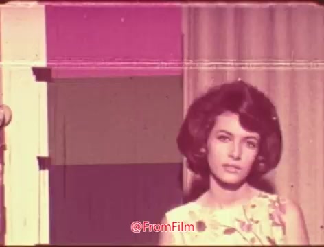 The still features a young woman with a stylish hairstyle, wearing a floral outfit. The colors are faded, typical of older 16mm film, lacking the vibrancy of processes like Technicolor. The background includes muted colors with a curtain and a vibrant pink wall, suggesting a candid or staged setting. The overall mood evokes a nostalgic atmosphere, characteristic of past eras.
