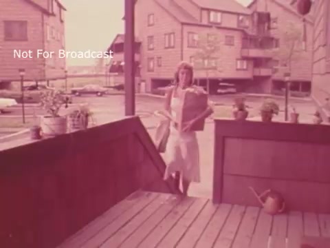The still shows a woman wearing a light-colored dress, carrying grocery bags as she steps onto a wooden porch. The scene is set in a residential area with multi-story buildings in the background and a few parked cars. The overall color tone is warm and vintage, indicative of the 1970s era. The context of the image relates to a PSA encouraging good nutrition, highlighting the importance of healthy eating, like incorporating bananas, even amidst busy lifestyles.