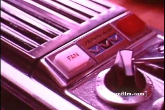 The still features a close-up of a Jenn Air grill control panel from a 1970s commercial. It displays a metallic design with a prominently labeled 'FAN' button and a round knob for temperature adjustment. The color scheme includes silver and white, reflecting the retro aesthetic of kitchen appliances from that era. The overall look conveys a vintage sophistication associated with home cooking technology of the time.