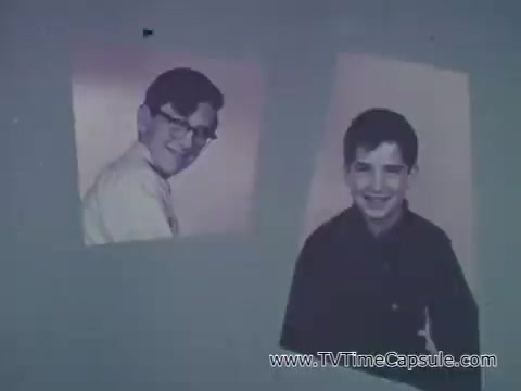 The still from the 'Camp Fire Girls' commercial features two boys' portraits. One boy with glasses smiles while facing the camera, portraying a sense of youthfulness and curiosity. The second boy has a more straightforward expression, presenting a friendly demeanor. The overall context emphasizes inclusion and diversity within the Camp Fire Girls organization, highlighting its appeal to both boys and girls in various roles. The setting reflects the 1960s era, aligning with the commercial's focus on community and teamwork in youth development activities.