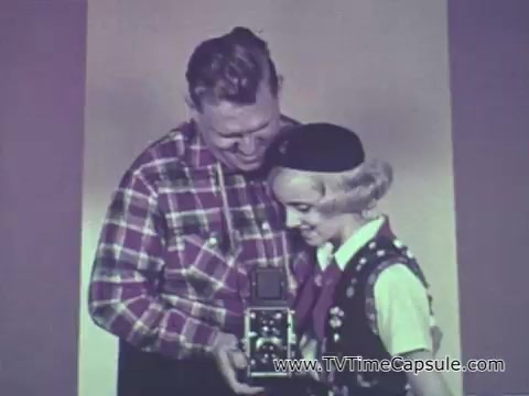 The still from the 1960s Camp Fire Girls commercial features a man and a young girl, both smiling warmly. The man, dressed in a plaid shirt, appears to be engaging with the girl, who is wearing a Camp Fire Girls vest and hat. She holds a camera, suggesting a moment of creativity or a fun activity. The scene reflects a friendly and supportive environment characteristic of the Camp Fire Girls program, which emphasizes community and leadership among youth.