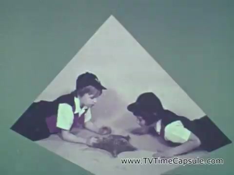 The still from the 1960s Camp Fire Girls commercial depicts two young children, likely dressed in uniforms, engaged in an activity on the ground. They seem to be focused on a shared object, possibly an art project or nature-related item, exhibiting curiosity and collaboration. The background is a simple, muted color, framing the scene in a diamond shape that directs attention to the children. The overall tone emphasizes community and learning, aligned with the values of the Camp Fire Girls organization as highlighted by Patrice Munsel in her introduction.