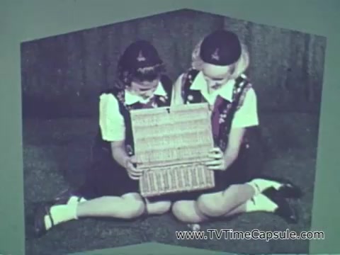 The still from the 1960s Camp Fire Girls commercial features two young girls sitting on the floor, dressed in similar outfits likely representing the Camp Fire Girls uniform. They are focused on a wicker basket or box that they are holding together. The setting appears casual, emphasizing a sense of community and engagement among the youth. The tone of the commercial aims to convey the inclusivity and various roles within the Camp Fire organization, highlighting both girls and boys, as well as adult leaders from different backgrounds.