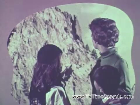 The still from the 'Camp Fire Girls' commercial features a group of girls and a woman, likely a leader or adult figure, looking towards a large, rocky backdrop. The scene suggests an outdoor or adventurous theme, highlighting the exploration aspects of the Camp Fire Girls program. The colors in the frame are likely muted or pastel, characteristic of the 1960s aesthetic. The setting emphasizes nature, possibly encouraging involvement in outdoor activities and teamwork. The overall vibe seems friendly and inviting, capturing the spirit of camaraderie and growth.