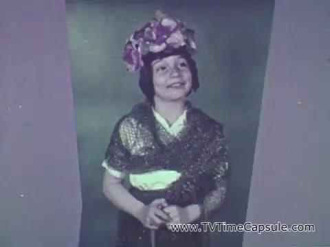 The 16mm still from the 'Camp Fire Girls' commercial features a young girl wearing a floral crown and a shawl. She is smiling, presenting a cheerful demeanor. The backdrop likely adds a colorful touch to the scene, aimed at showcasing the diversity of participants in the Camp Fire Girls program. The context emphasizes the inclusivity of the program, highlighting involvement from various community members, including girls, boys, and adult leaders.