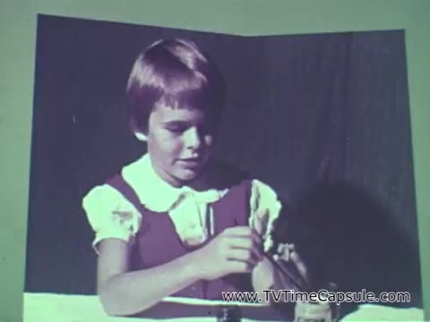 The still from the 1960s Camp Fire Girls commercial features a young girl with short hair, wearing a white collared shirt under a dress. She is focused on a task, likely involving crafts or activities, typical of what Camp Fire Girls promoted. The background appears simple, emphasizing her engagement in the activity. The commercial aims to highlight the diverse involvement of girls and boys, along with leaders from different backgrounds, in the Camp Fire Girls program.