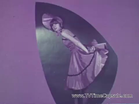The still from the 'Camp Fire Girls' commercial features a young woman dressed in a playful outfit, characterized by a pink dress with ruffled edges and a matching hat. She poses in front of a vibrant purple backdrop, accentuating the lively and whimsical tone of the commercial. The scene likely aims to convey the fun and inclusive spirit of the Camp Fire Girls organization, emphasizing its appeal to various youth and adult demographics.