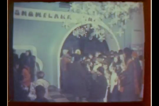 The still depicts a bustling scene inside a department store, likely during the holiday season. Children and adults gather around a festive entrance, decorated with lights and a whimsical arch. The atmosphere is lively, emphasizing the excitement of visiting Santa. The overall tone reflects a nostalgic, cheerful holiday spirit typical of the 1970s.